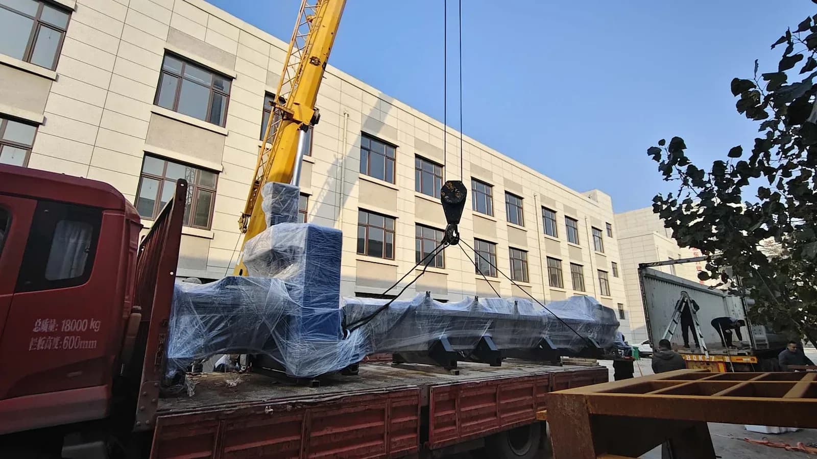 Кран поднимает с борта грузовика станок, обёрнутый плёнкой, на фоне заводского здания
