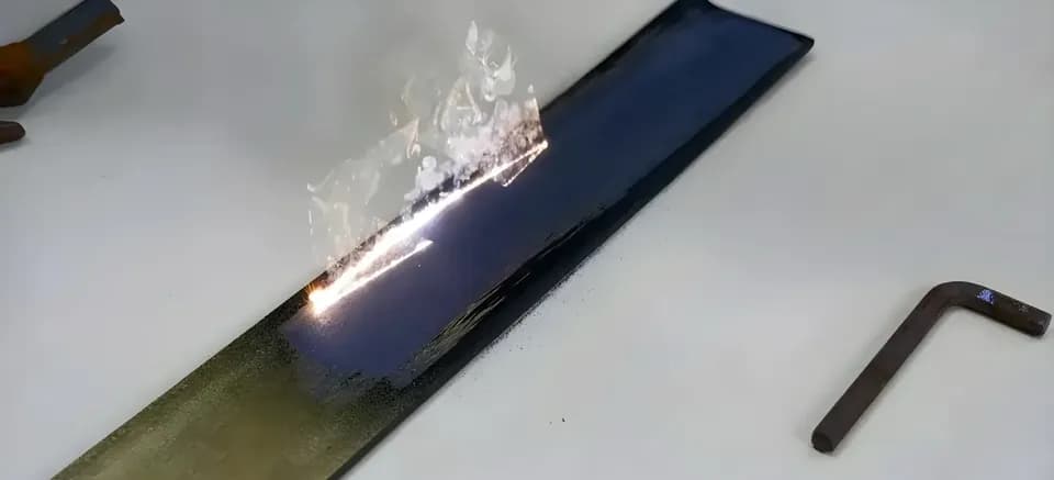 Полоса чёрного пластика на белом столе горит, над поверхностью поднимаются дым и вспышка пламени